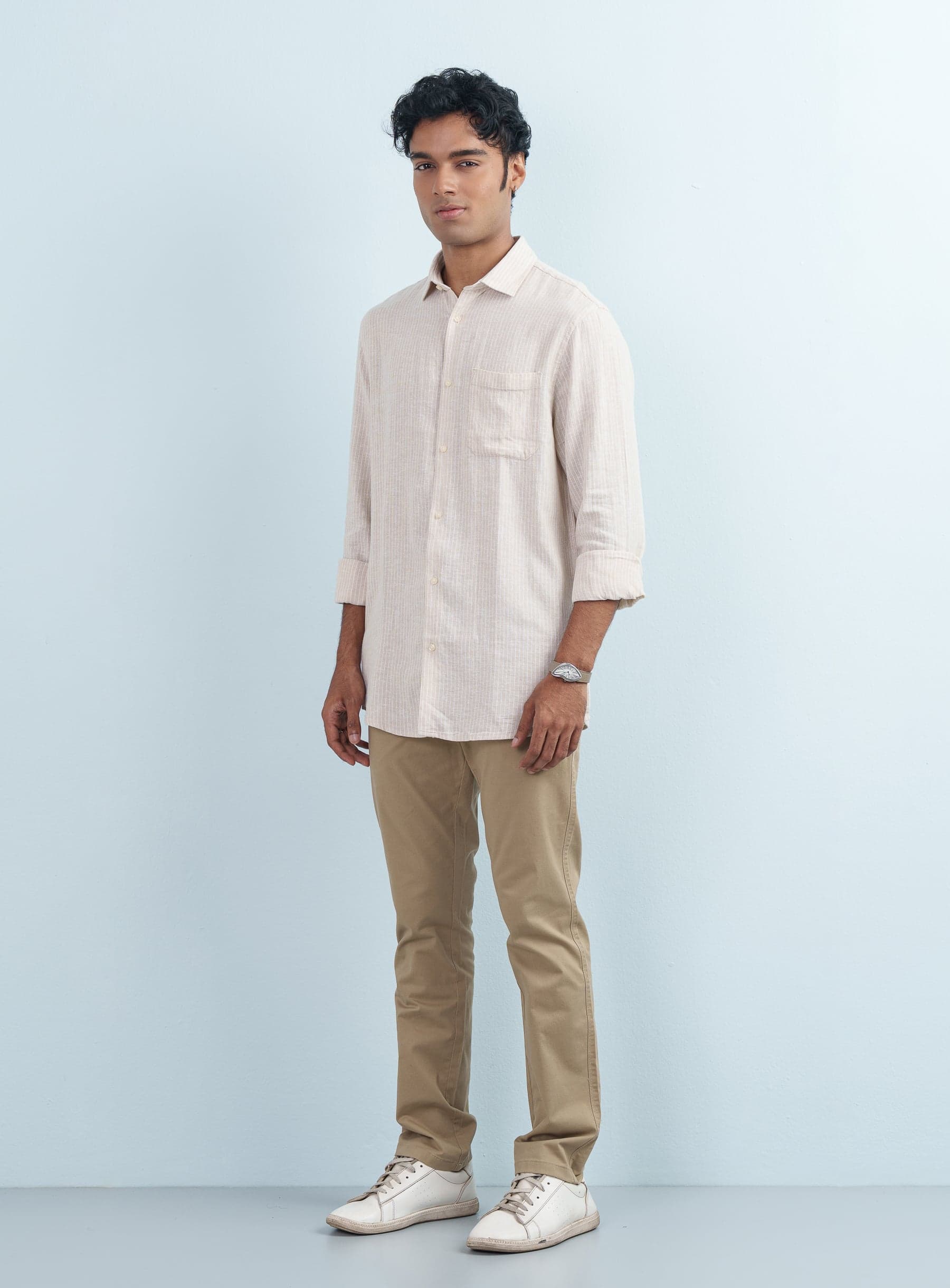 Full-length studio shot of a man wearing a beige striped cotton mixed casual shirt full sleeve with khaki trousers and white sneakers.