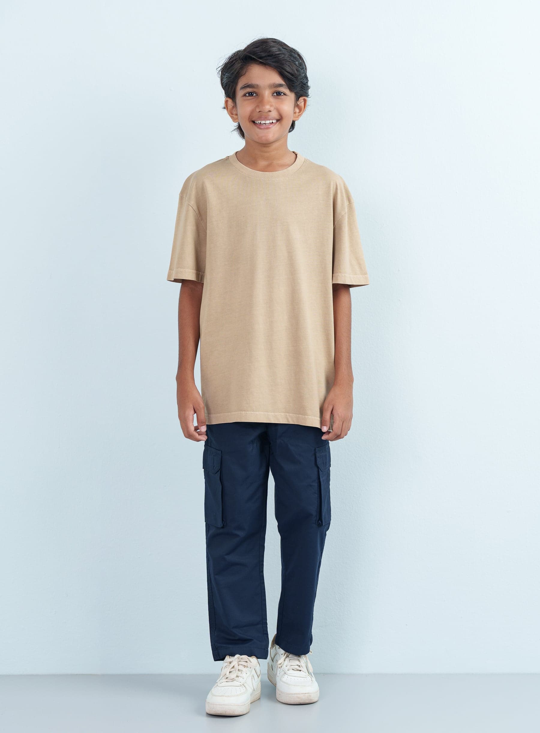 Full-length front shot of a smiling boy modeling a casual beige T Shirt Half with an oversized fit paired with navy blue cargo pants.