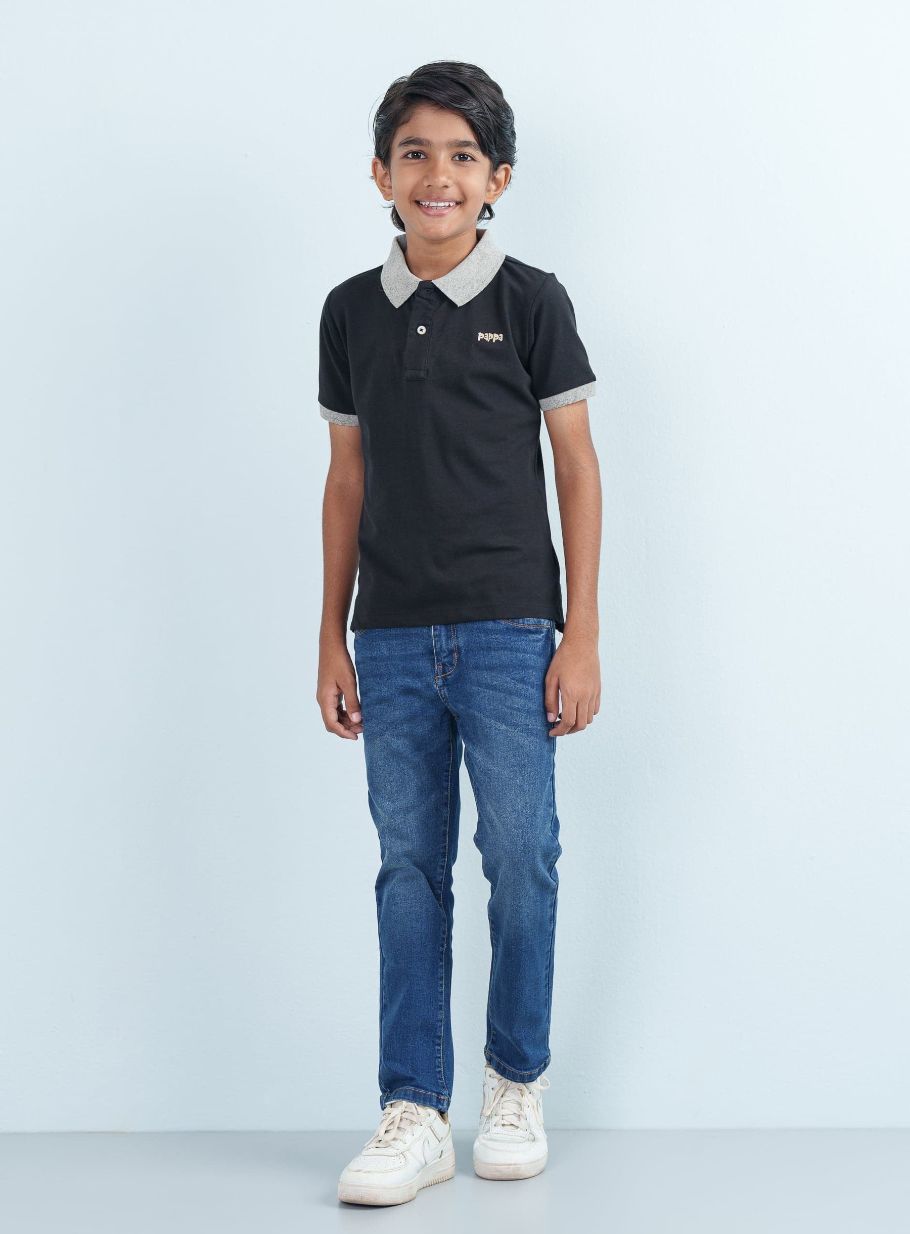 Full-length front shot of a young boy modeling a black Polo Half paired with classic blue denim jeans and white sneakers.