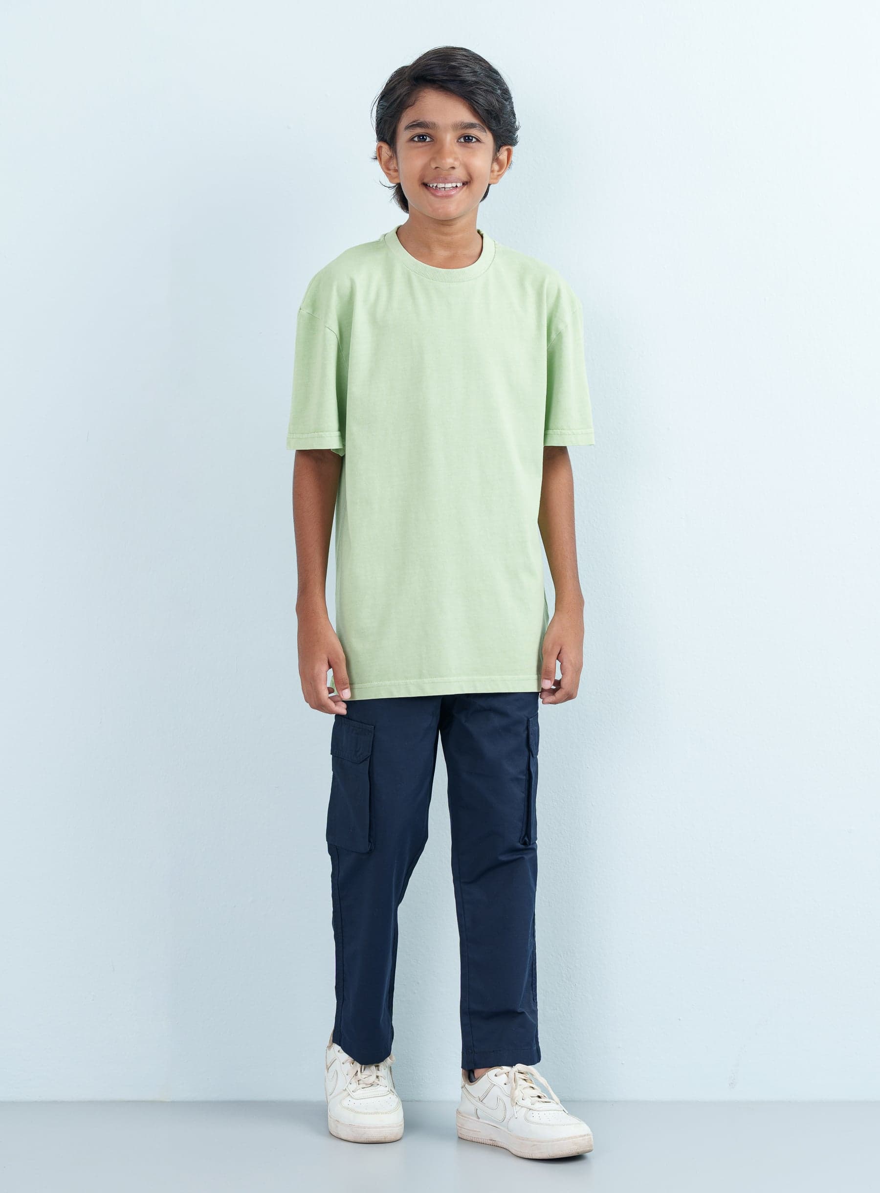 Full-length front shot of a smiling boy modeling a casual mint green T Shirt Half with an oversized fit paired with navy blue cargo pants.