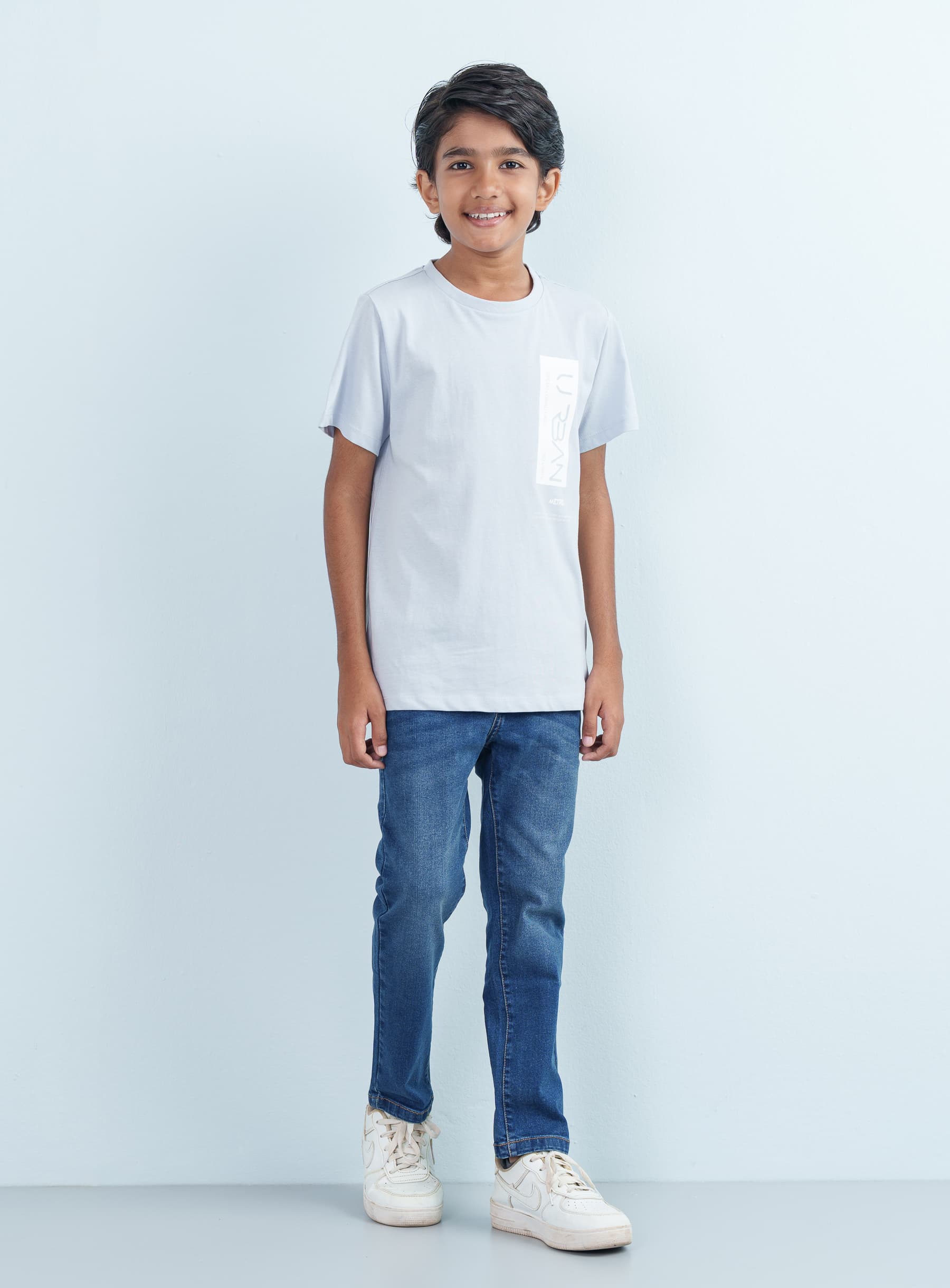 Full-length studio shot of a smiling young boy wearing a light gray cotton T Shirt Half with an urban chest graphic paired with blue jeans.