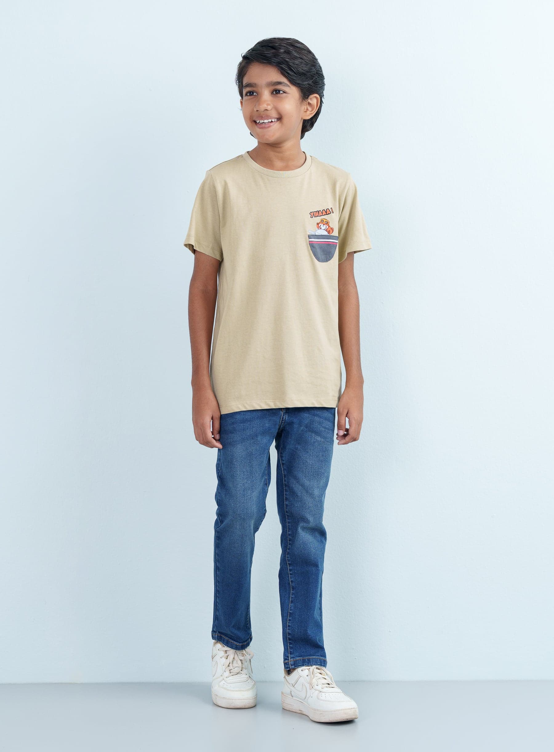 Full-length front shot of a smiling boy modeling a casual khaki T Shirt Half with a unique pocket graphic paired with classic blue jeans.