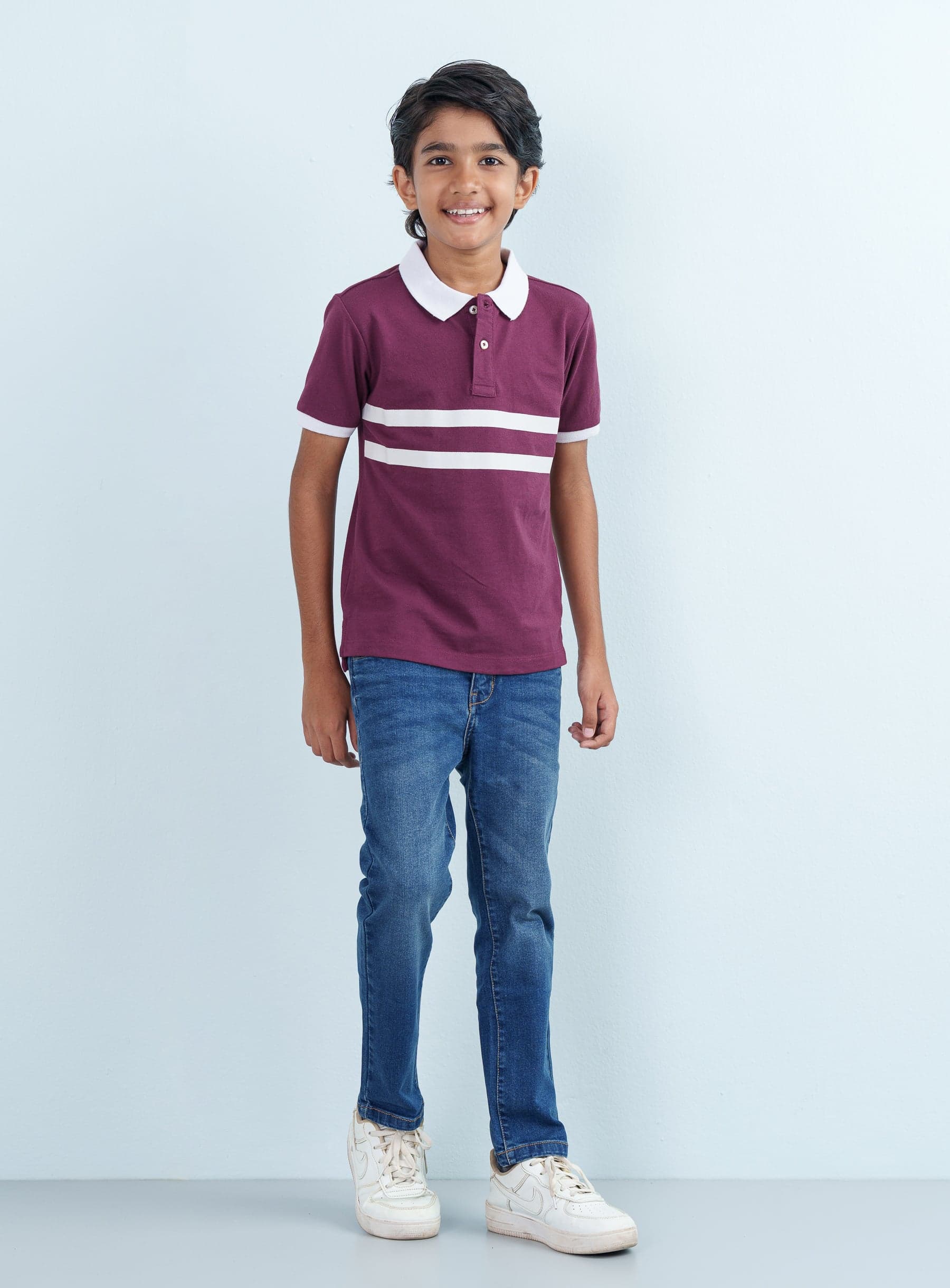 Full-length front shot of a smiling young boy modeling a maroon Polo Half with dual white stripes paired with classic blue jeans.