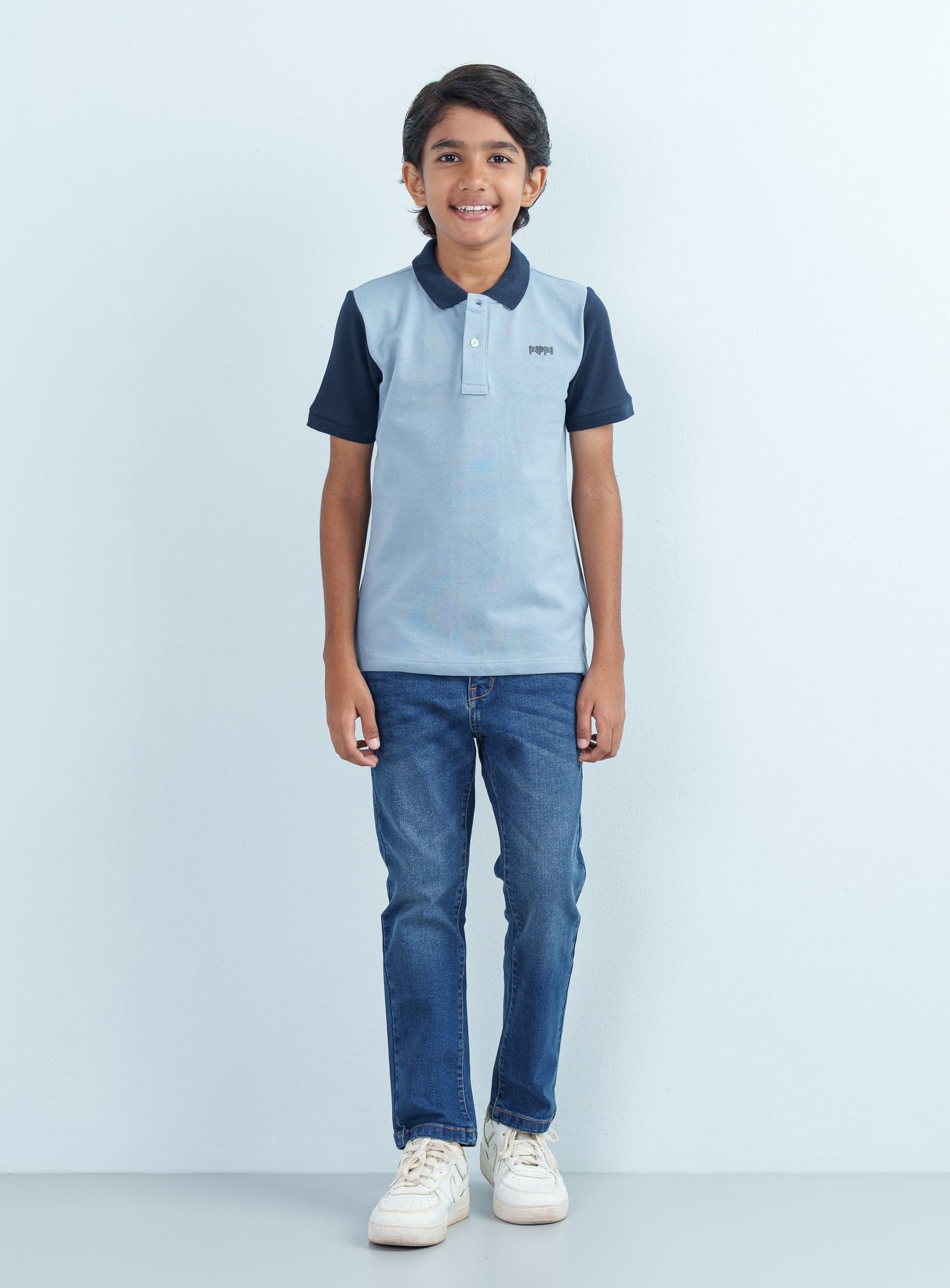 Full-length front shot of a young boy wearing a light blue and navy Polo Half styled with classic blue jeans and white sneakers.