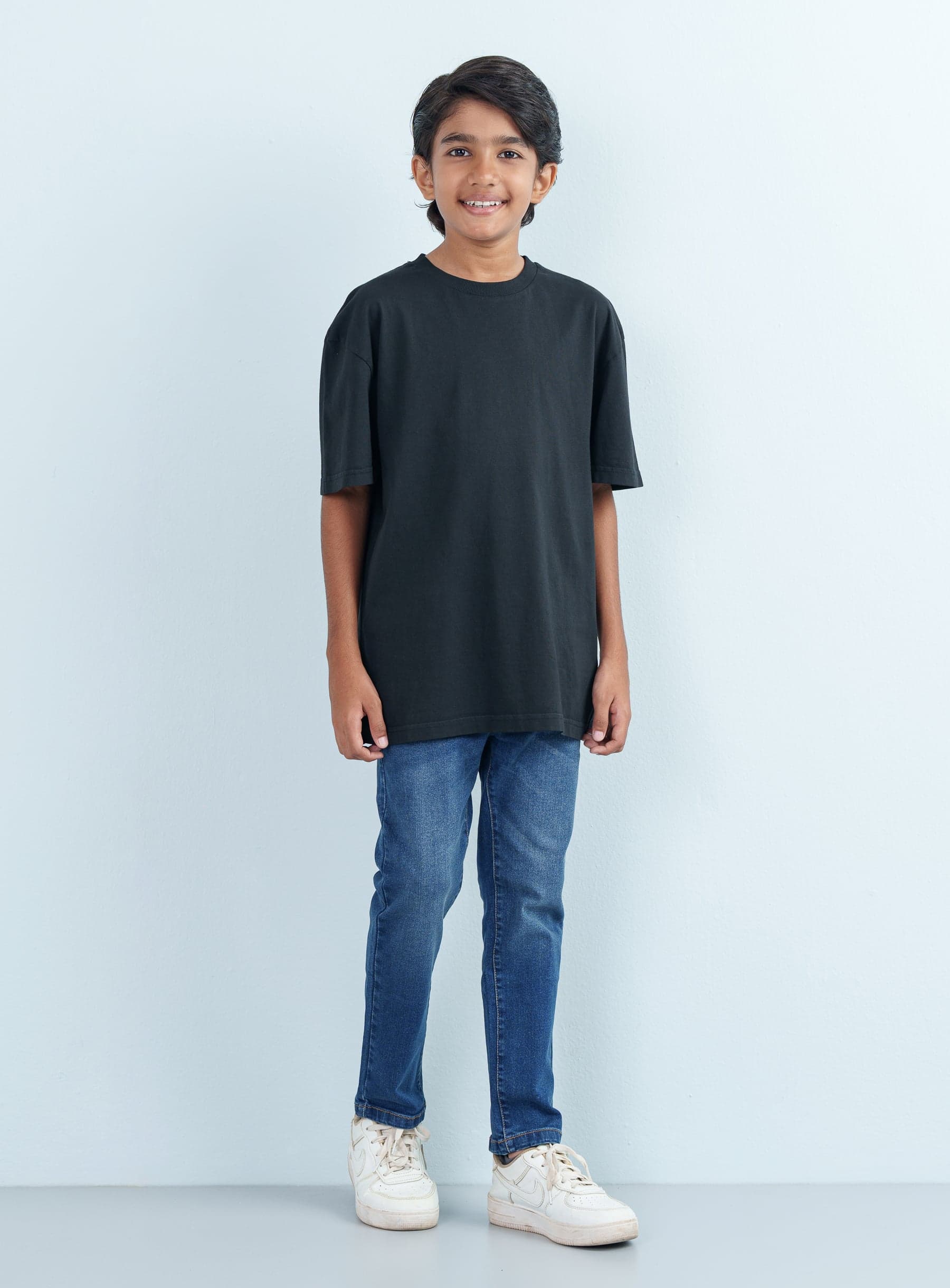 Full-length front shot of a smiling boy modeling a casual black T Shirt Half with an oversized fit paired with classic blue jeans.